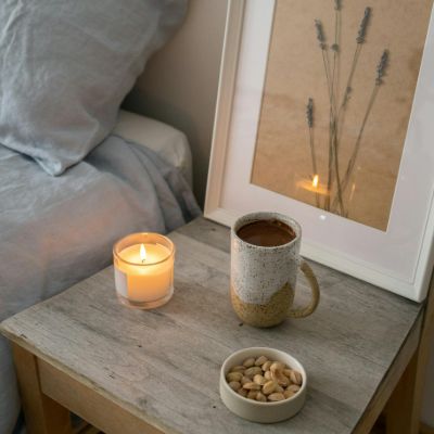 A cup of bedtime tea alongside a lit candle besides a warm bed in the morning. 