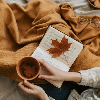 A book with a dried maple leaf and a cup of fall tea being held by a woman. 